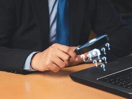 A businessman is holding a magnifying glass and looking at a question mark symbol on a laptop while sitting in the office photo