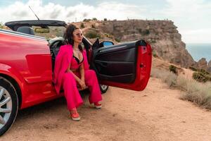 A woman in a pink suit sits in the open door of a red convertible. The scene is set on a rocky hillside overlooking the ocean. The woman is posing for a photo. photo