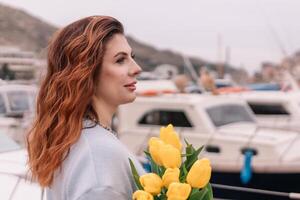 Woman holds yellow tulips in harbor with boats docked in the background., overcast day, yellow sweater, mountains photo