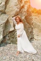 A woman in a white dress is standing on a rocky beach. Concept of serenity and tranquility, as the woman is enjoying the natural beauty of the beach. photo