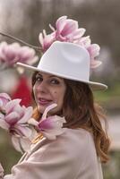 Woman magnolia flowers, surrounded by blossoming trees., hair down, white hat, wearing a light coat. Captured during spring, showcasing natural beauty and seasonal change. photo