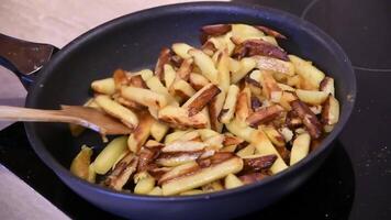 Hand with wooden paddle stir homemade French fries in frying pan at kitchen stove. Cooking of crispy fried potatoes with curry. Using frying spatula to mix hot dish. Slow motion video