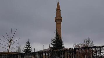 Mosque minaret and trees in cold weather with falling snow. Islamic architecture of brick masonry minaret tower on grey cloudy sky background. Snowflakes fly in air. Travel Turkey in winter video
