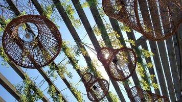 Wicker vine lamp shades hang and sway under wooden roof of arbor. Sun flare shine through vine woven lamp chandelier shades with retro light bulbs inside and tangled lianas. Slow motion low angle view video