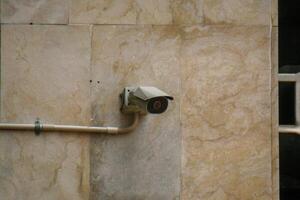 A security camera mounted on a beige marble wall, connected by a conduit cable, symbolizing surveillance, safety, and modern security systems. photo