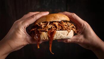 close-up of hands gripping a pulled pork sandwich with barbecue sauce oozing from the sides against a dark rustic background photo