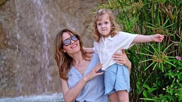 Mother holding young daughter by waterfall, child raises hand in greeting, looking at camera, both smiling, enjoying bonding moment in nature, carefree family time, concept of joyful connection video