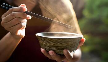close-up of hands holding a steaming bowl of soup with chopsticks, thick steam rising in soft natural light photo
