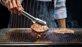 close-up of a chef grilling meat with tongs over a sizzling hot grill, showcasing smoky flames and seared steak in a professional kitchen setting photo