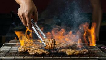 close-up of hands using tongs to grill juicy meat over open flames, with smoke rising on a barbecue grill photo