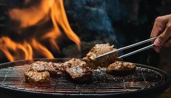 close-up of hands using tongs to grill sizzling pieces of meat over open flames with smoke rising on a barbecue grill photo
