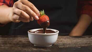 close-up of hands dipping fresh strawberries into melted chocolate, creating a rich dessert treat on a wooden table photo