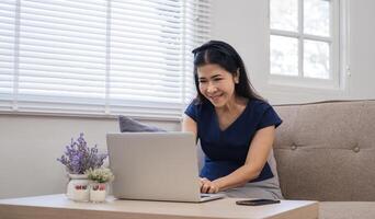 Happy Asian retired woman in her 60's enjoying social media on laptop while relaxing in living room. photo