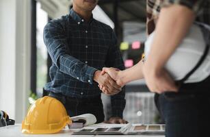 Two Male Engineers Shaking Hands in Modern Office Environment, Collaborating on Construction Project with Safety Helmet and Design Plans on Desk photo