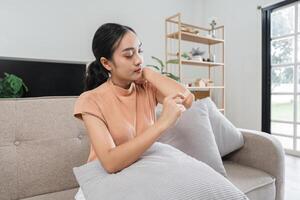Young Woman with Injured Elbow Sitting on Sofa at Home, Holding and Examining Her Sore Arm, Experiencing Pain and Discomfort, Indoor Casual Setting, Health and Wellness Concept photo