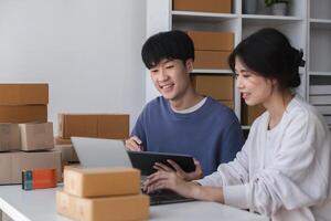 Young Couple Managing Their Online Small Business from Home, Surrounded by Shipping Boxes and Working on a Laptop and Tablet in a Modern Home Office Setting photo