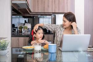 Asian mother working on laptop while interacting with daughter in kitchen. Concept of balancing work and family time photo