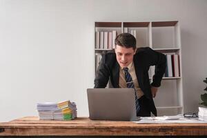 Businessman Working in Modern Office with Laptop and Documents on Desk, Professional Corporate Environment, Focused and Productive Work Atmosphere, Office Interior with Shelves and Books photo