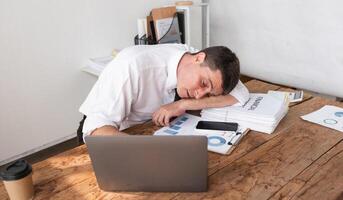 Businessman exhausted and asleep at desk with laptop and financial reports, concept of workplace fatigue and stress photo