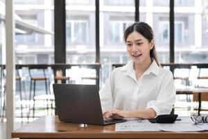 Professional Business Woman Working on Laptop in Modern Office Setting with Documents and Calculator on Desk, Smiling and Engaged in Productive Work, Large Windows in Background photo