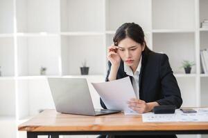 Stressed Business Woman Working at Desk with Laptop and Documents in Modern Office Environment, Experiencing Workload Pressure and Deadlines, Wearing Formal Attire, Concentrating on Paperwork photo