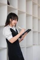 Young Female University Student Taking Notes on Digital Tablet in Modern Library Setting, Focused on Learning and Education, Wearing Casual Outfit, Engaged in Academic Activities, Smiling photo