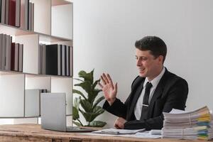 Businessman Working in Modern Office Environment, Engaging in Call, Professional Attire, Office Desk with Documents, Bookshelf in Background, Daylight Setting, Corporate Workspace photo