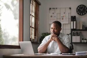 African American businessman feeling stressed while working using laptop Take notes on documents in the office and prepare work reports. photo