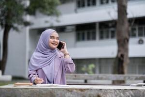 Confident Muslim Girl Student in Hijab Using Smartphone and Studying Outdoors at University Campus photo