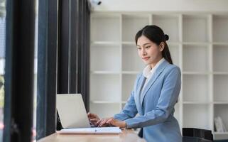 Professional Business Woman Working on Laptop in Modern Office Setting with Natural Light and Minimalist Design, Focused and Productive, Wearing Formal Attire, Engaged in Corporate Tasks photo