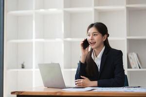 Professional Business Woman Working in Modern Office Environment, Engaged in Phone Call and Using Laptop, Dressed in Formal Attire, Demonstrating Productivity and Professionalism photo