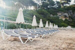 Scenic view in sunny morning to a chaise lounges and umbrellas on the sandy beach without people,sea and rocks on the background.Close up. High quality photo