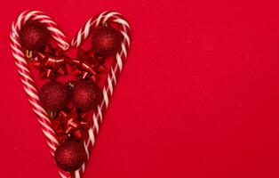 Top view photo heart lined with Christmas lollipops and decorated with Christmas tree toys of red colour,on red background for greeting card. Xmas backdrop with space for text.Copyspace.
