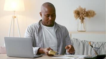 African American man in his 30s sits at a desk in a stylish home office. He is counting cash while focused on financial matters, embodying a contemporary lifestyle. video