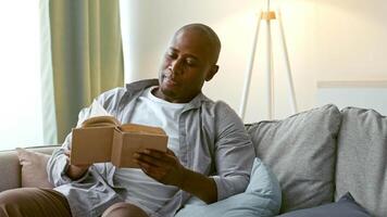 A man in his 30s enjoys reading a book on a comfortable couch in a bright, modern living room. The atmosphere is calm, promoting relaxation and leisure time. video