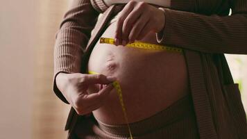 An expectant mother measures her growing baby bump using a tape measure while seated comfortably at home. She smiles, reflecting the joy of pregnancy. video