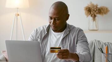African American man sits comfortably at home, focused on his laptop while holding a credit card. He is engaged in online shopping, enjoying the relaxed atmosphere. video