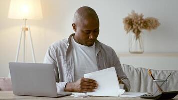 A focused African American man in his 30s reviews paperwork while sitting at a desk with a laptop in a cozy home office. Soft light adds to the peaceful atmosphere of productivity. video