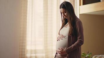 A pregnant woman stands in a warmly lit room, gently touching her belly with a serene expression. She is surrounded by natural light and a tranquil atmosphere, creating a peaceful moment. video
