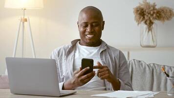 African American man in his 30s is sitting at a home workspace, engaged with his smartphone. He is casually dressed and appears focused on his device, surrounded by a cozy environment. video