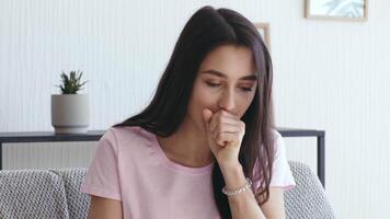 A young woman sits on a couch in her home, visibly unwell, coughing as she deals with the symptoms of the flu or a respiratory illness while in isolation. video