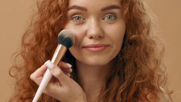 A cheerful young woman with vibrant curly red hair stands confidently, smiling. She appears relaxed and approachable, set against a neutral beige backdrop, creating a warm atmosphere. video