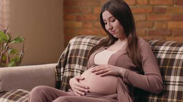 A pregnant woman sits comfortably on a couch, gently cradling her belly with a look of contentment. Natural light filters through the window, creating a warm atmosphere. video