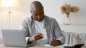 A man in his 30s is focused on writing notes while reviewing a document in his comfortable home office. He sits at a wooden desk with a laptop and a plant nearby, creating a productive atmosphere. video