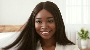 A cheerful African American woman with long, smooth hair sits in a cozy, well-lit space, holding a wooden brush and smiling warmly towards the viewer. She appears relaxed and happy. video