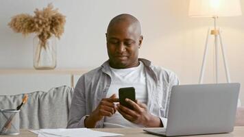 A African American man in his 30s is seated comfortably at home, concentrating on his smartphone while a laptop is open nearby. Papers are scattered around, indicating he is multitasking. video