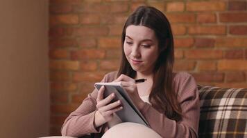 A young pregnant woman sits comfortably on a couch, writing in a notebook with a focused expression. The warm brick wall and soft lighting create a cozy and inviting atmosphere. video