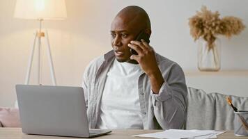African American man in his 30s is engaged in a phone conversation while seated at a desk. He appears focused and relaxed, surrounded by a cozy home setting with warm lighting. video