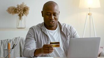 A man in his 30s smiles while holding a credit card in front of his laptop at home. He is engaged in online shopping, enjoying a relaxed moment in his living space. video