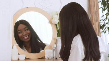 African American woman smiles while brushing her long hair in front of a round mirror. The bright bathroom features elegant decor and nature-inspired elements, creating a refreshing atmosphere. video